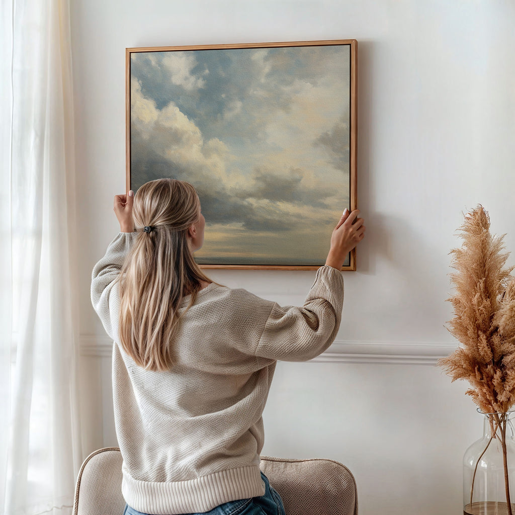 A woman with blonde hair in a ponytail hangs the Heavenly Drift Canvas Art, featuring a cloudy sky, on a white wall in a serene living room with a beige chair and dried pampas grass for cozy, vintage decor.
