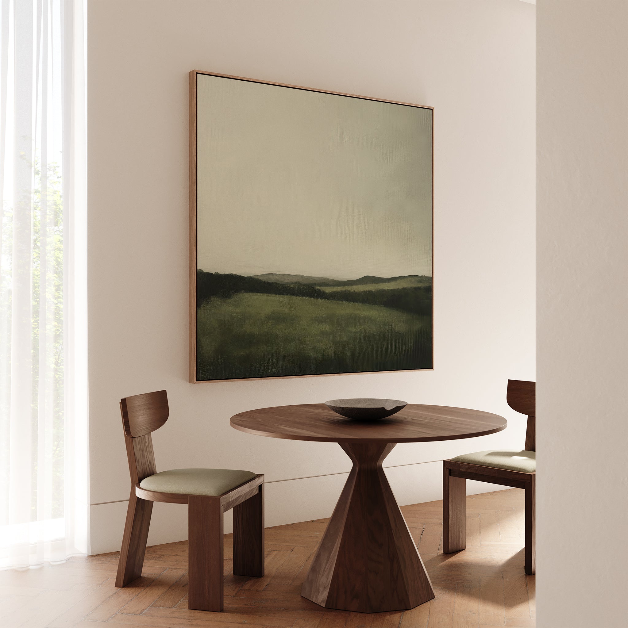 A minimalist dining area features a round wooden table, two cushioned chairs, a ceramic bowl, and the Hills at Dusk Canvas Art on a light wall beside a window with sheer curtains.