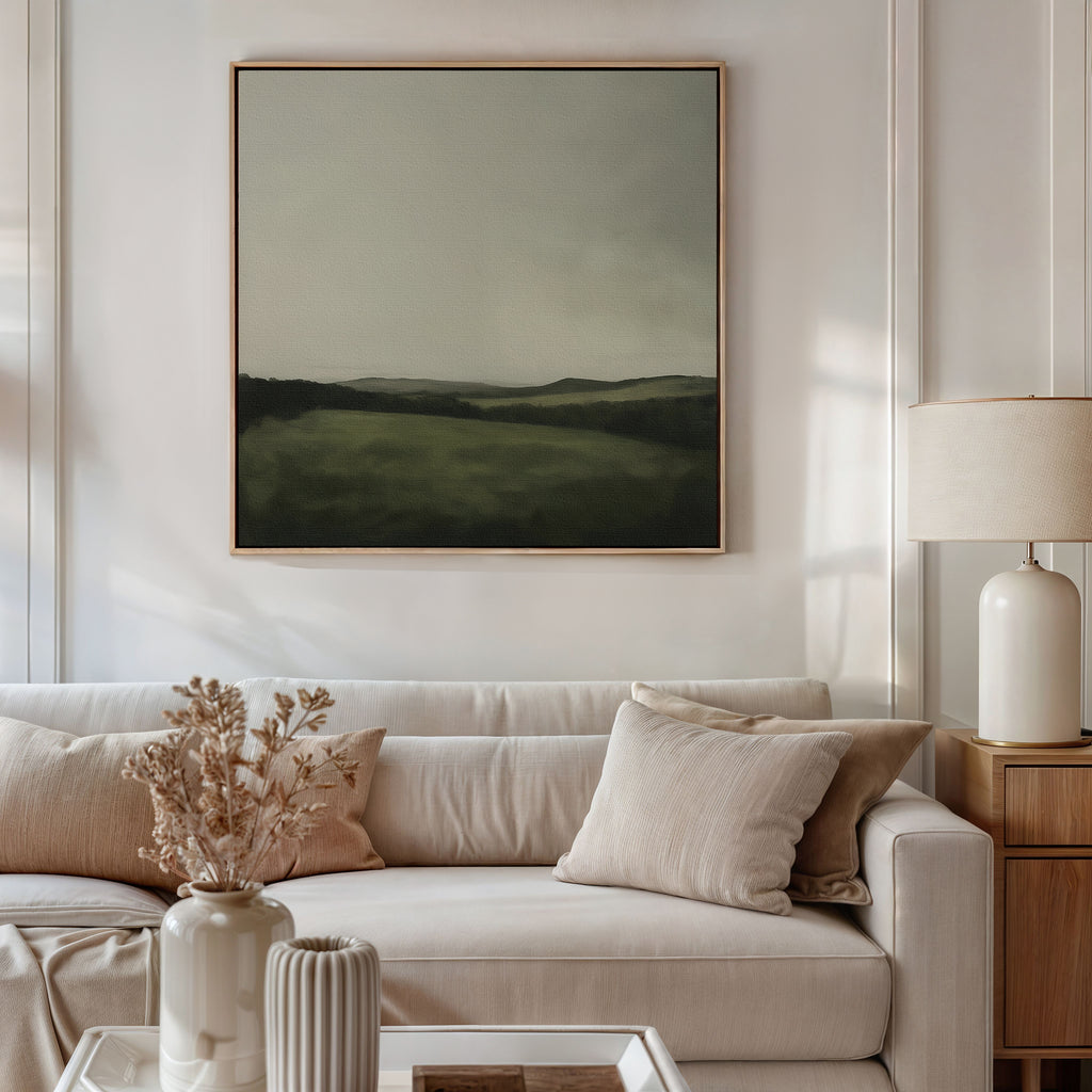 A modern living room features a light beige sofa with cream pillows, a dried floral arrangement in a vase, a table lamp, and the Hills at Dusk Canvas Art—a minimalist wall print of a misty field and hills—displayed above the sofa.
