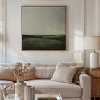 A modern living room features a light beige sofa with cream pillows, a dried floral arrangement in a vase, a table lamp, and the Hills at Dusk Canvas Art—a minimalist wall print of a misty field and hills—displayed above the sofa.