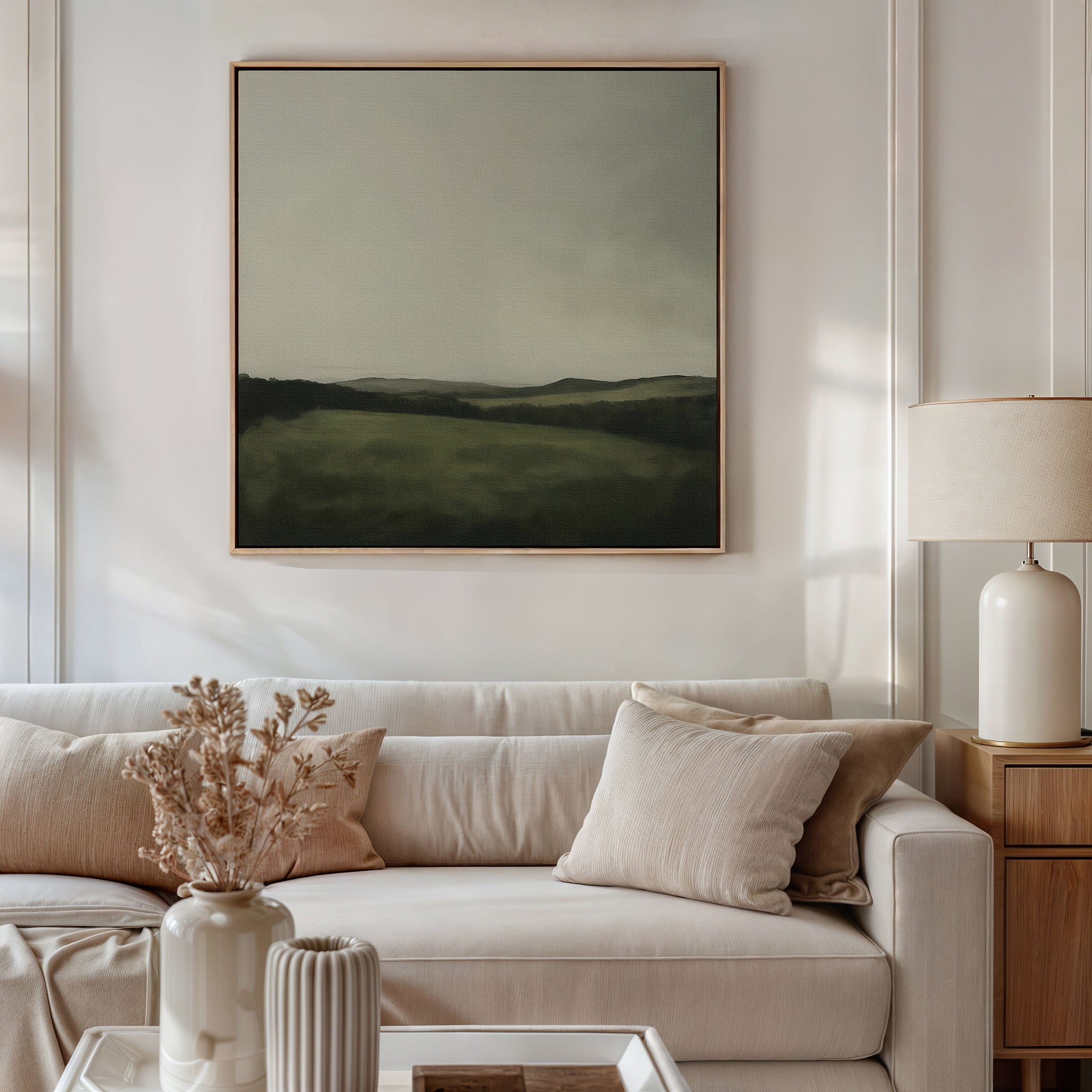 A modern living room features a light beige sofa with cream pillows, a dried floral arrangement in a vase, a table lamp, and the Hills at Dusk Canvas Art—a minimalist wall print of a misty field and hills—displayed above the sofa.