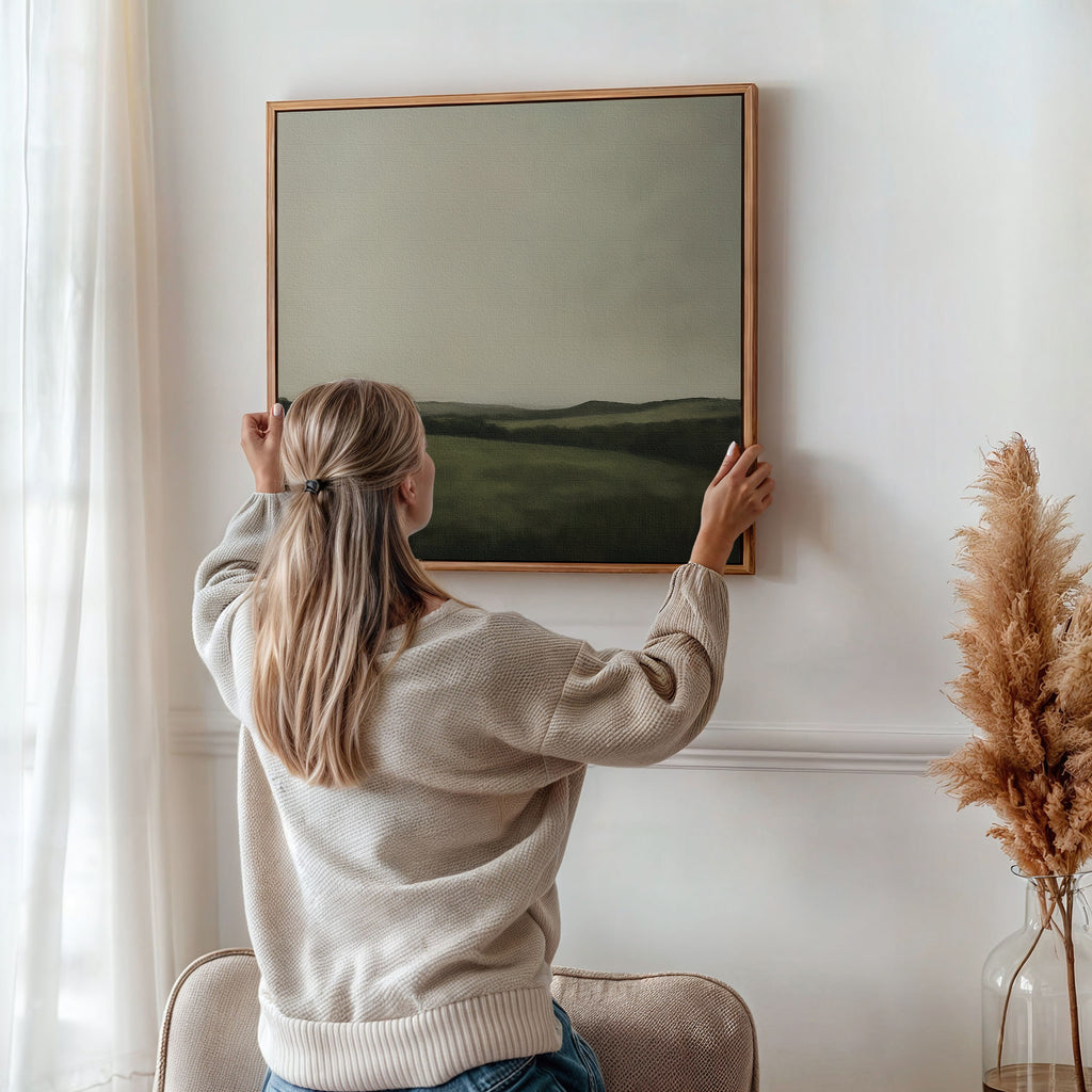 A woman with long blonde hair hangs the Hills at Dusk Canvas Art on a white wall in a bright, cozy room decorated with neutral tones and pampas grass in a vase.