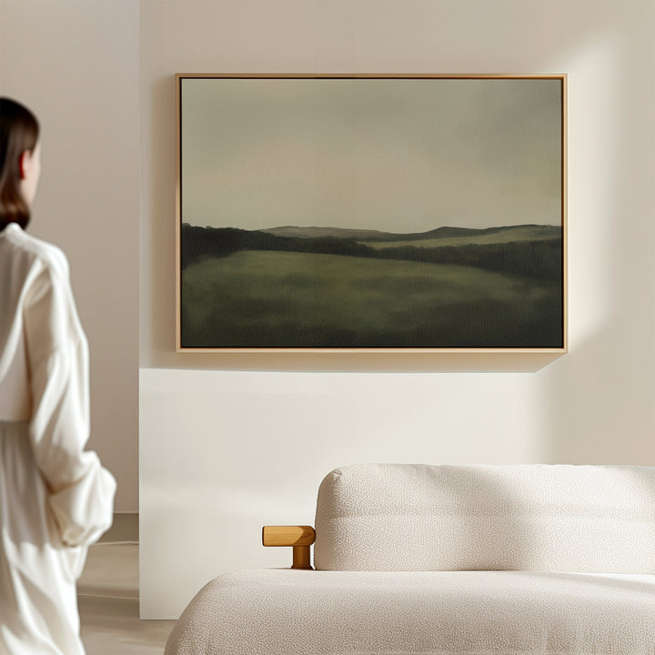 A person in a white shirt stands in a minimalist room, admiring the Hills at Dusk Canvas Art – Horizontal above a light-colored sofa, adding an elegant touch to the living room decor.