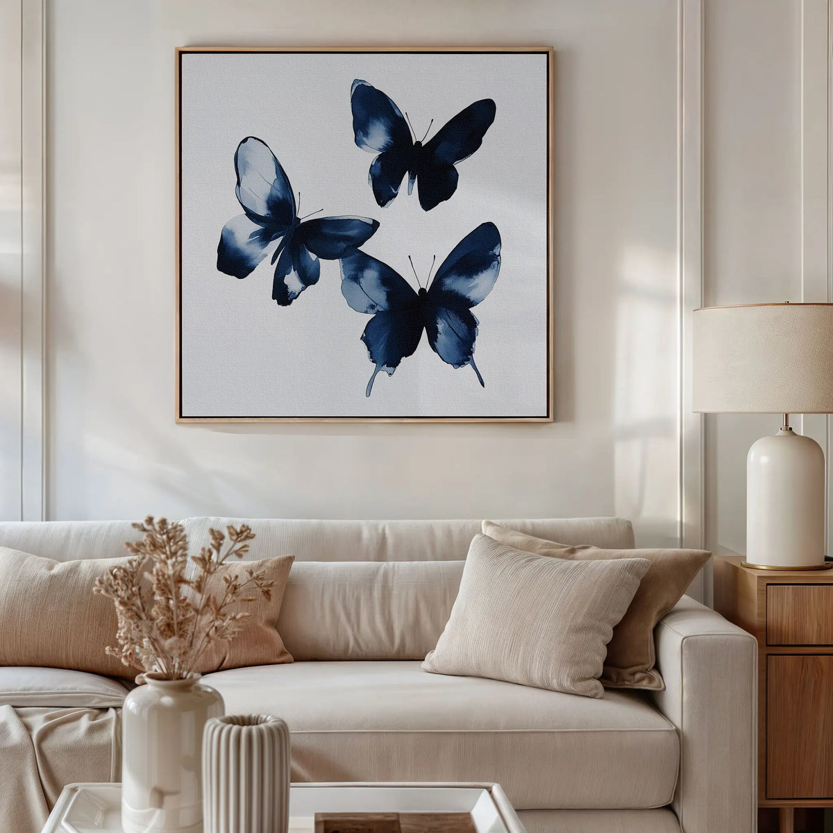 A living room features a white sofa, beige cushions, wooden side table with lamp, and the Indigo Flutter Canvas Art. A vase with dried flowers on the coffee table completes the modern home decor charm.