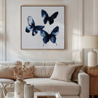 A living room features a white sofa, beige cushions, wooden side table with lamp, and the Indigo Flutter Canvas Art. A vase with dried flowers on the coffee table completes the modern home decor charm.