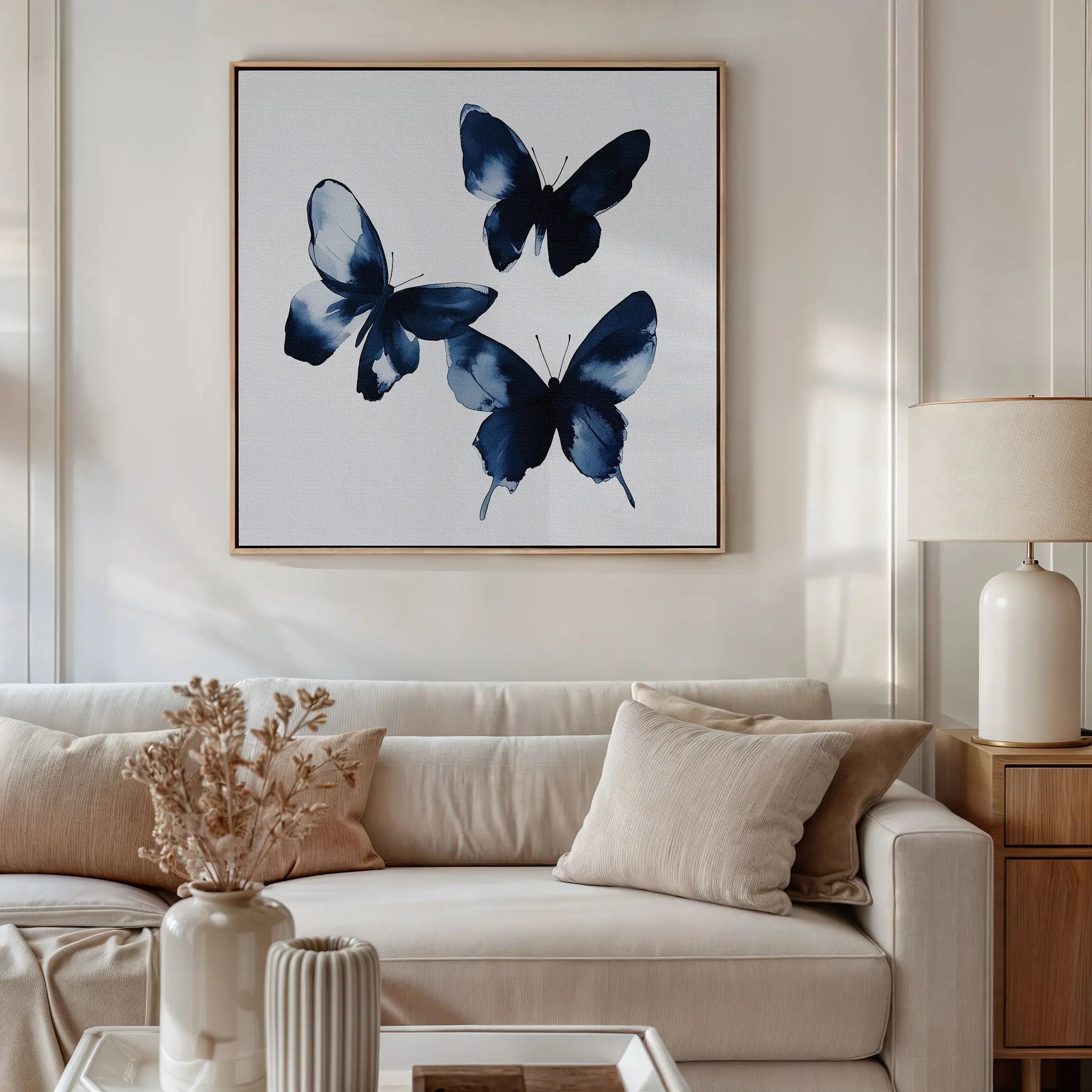 A living room features a white sofa, beige cushions, wooden side table with lamp, and the Indigo Flutter Canvas Art. A vase with dried flowers on the coffee table completes the modern home decor charm.
