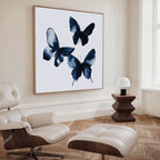 A modern living room with a white lounge chair and ottoman, wooden side table with a lamp, and a large Indigo Flutter Canvas Art featuring three abstract blue butterflies on a white background.