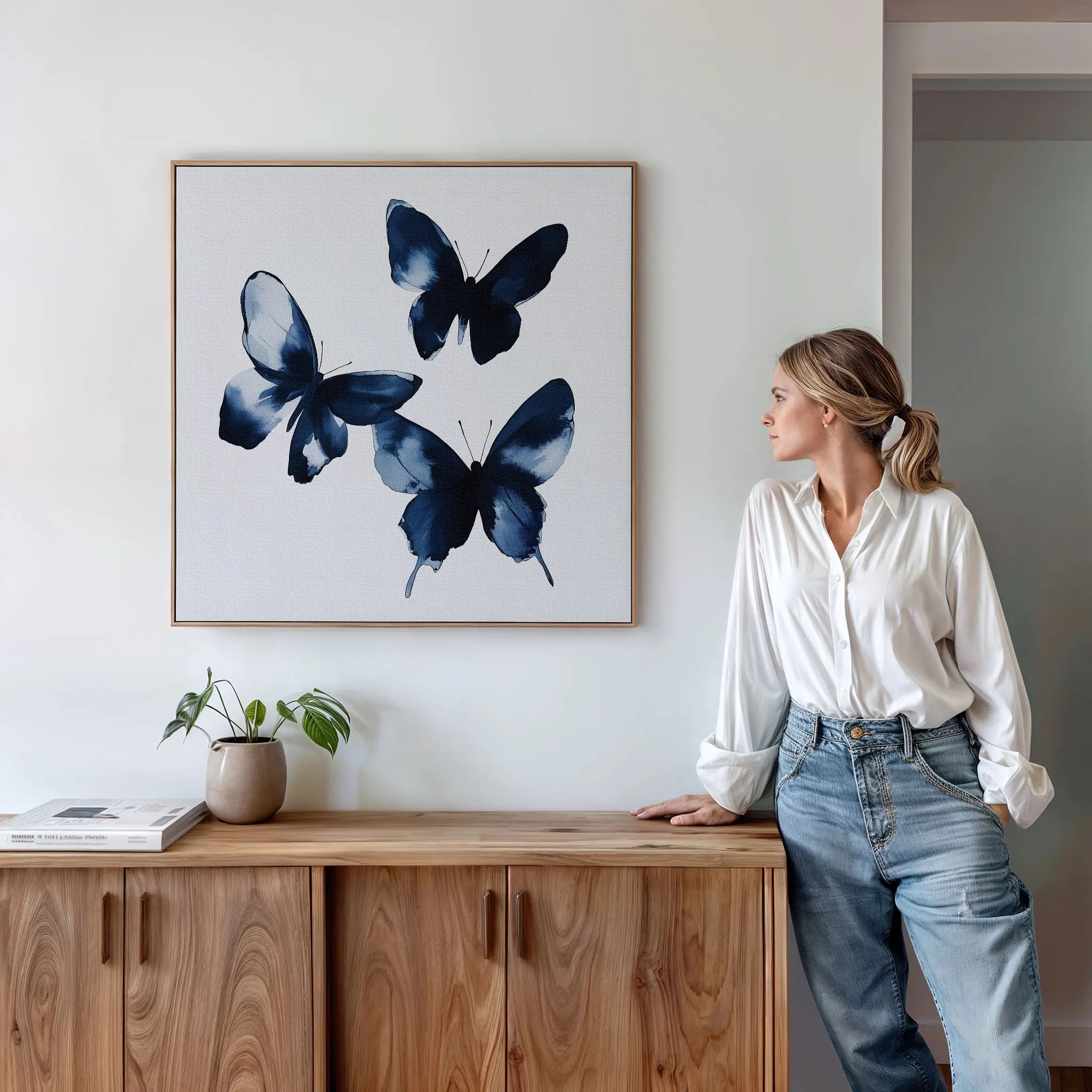A woman in a white blouse and jeans stands by a wooden cabinet, admiring the Indigo Flutter Canvas Art. A potted plant and book enhance the modern home decor on the cabinet against the white wall.