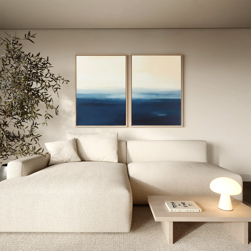 A minimalist living room features a light beige sectional sofa, a low wooden coffee table with a book and round lamp, an indoor plant, and the Indigo Horizon Set of 2 wall art—perfect for modern living room decor.