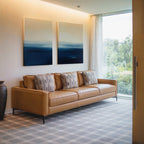 A modern living room with a tan leather sofa, four patterned cushions, the Indigo Horizon Set of 2 wall art above, a large window with sheer curtains, and a decorative vase on the floor—ideal for modern living room decor.