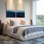A modern bedroom features a large bed with white and gray bedding, several white pillows, Indigo Horizon Set of 2 wall art above the headboard, a large window letting in natural light, and decorative vases on each side.