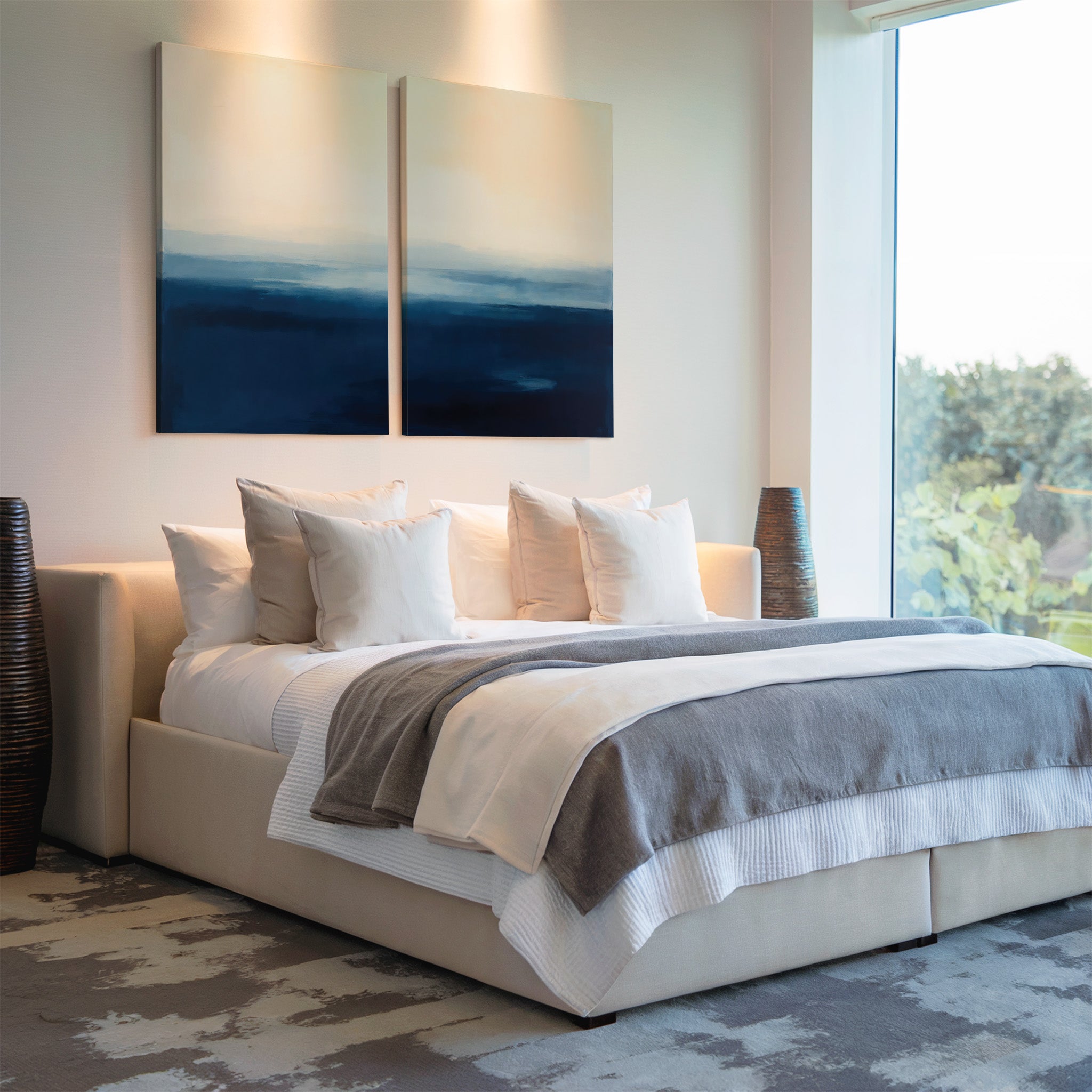 A modern bedroom features a large bed with white and gray bedding, several white pillows, Indigo Horizon Set of 2 wall art above the headboard, a large window letting in natural light, and decorative vases on each side.