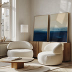 A modern living room with two rounded white armchairs, a low wooden coffee table, and the Indigo Horizon Set of 2 wall art leaning on a wooden sideboard beneath large windows—perfect for modern decor.