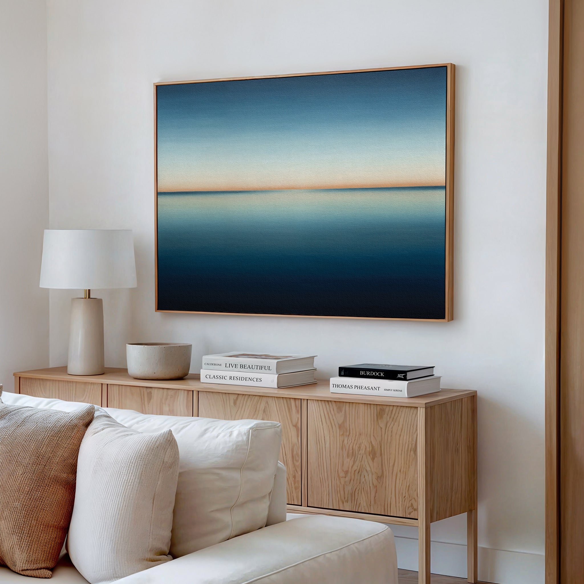 A modern living room with a wooden sideboard, white lamp, books, a bowl, and a beige sofa features the Indigo Passage Canvas Art – Horizontal above the sideboard, lending a minimalist and serene touch to the space.