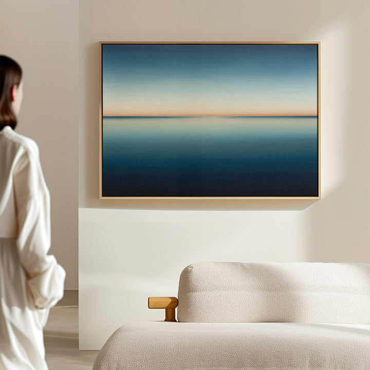 A woman in white stands in a bright, minimal room, admiring the Indigo Passage Canvas Art – Horizontal with blue and beige tones above a cream sofa—perfect for modern living room decor.