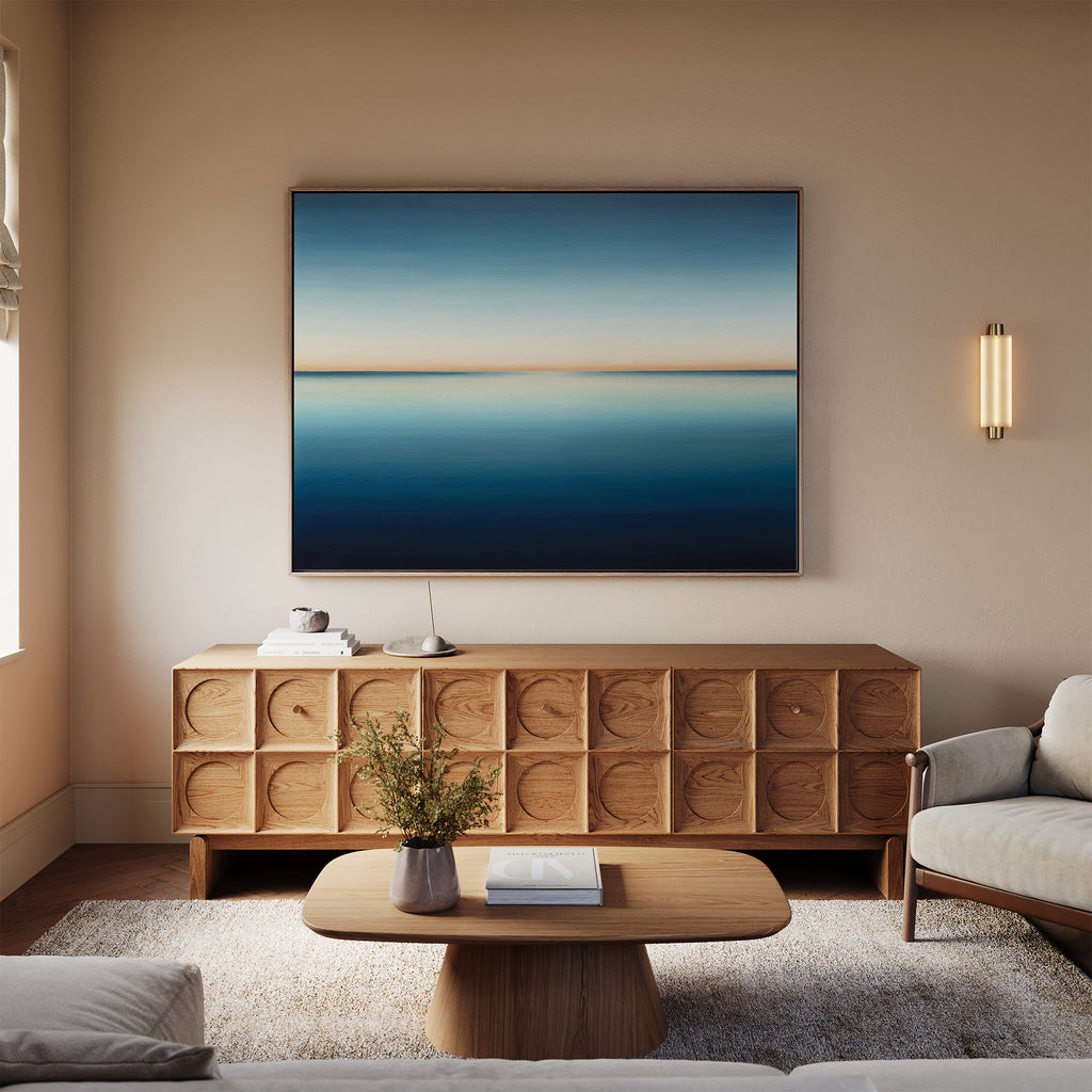 A modern living room features a wooden sideboard, the Indigo Passage Canvas Art – Horizontal in blue and beige tones, a coffee table with books, a light rug, beige sofa, wall sconce, and a small vase with flowers for stylish decor.