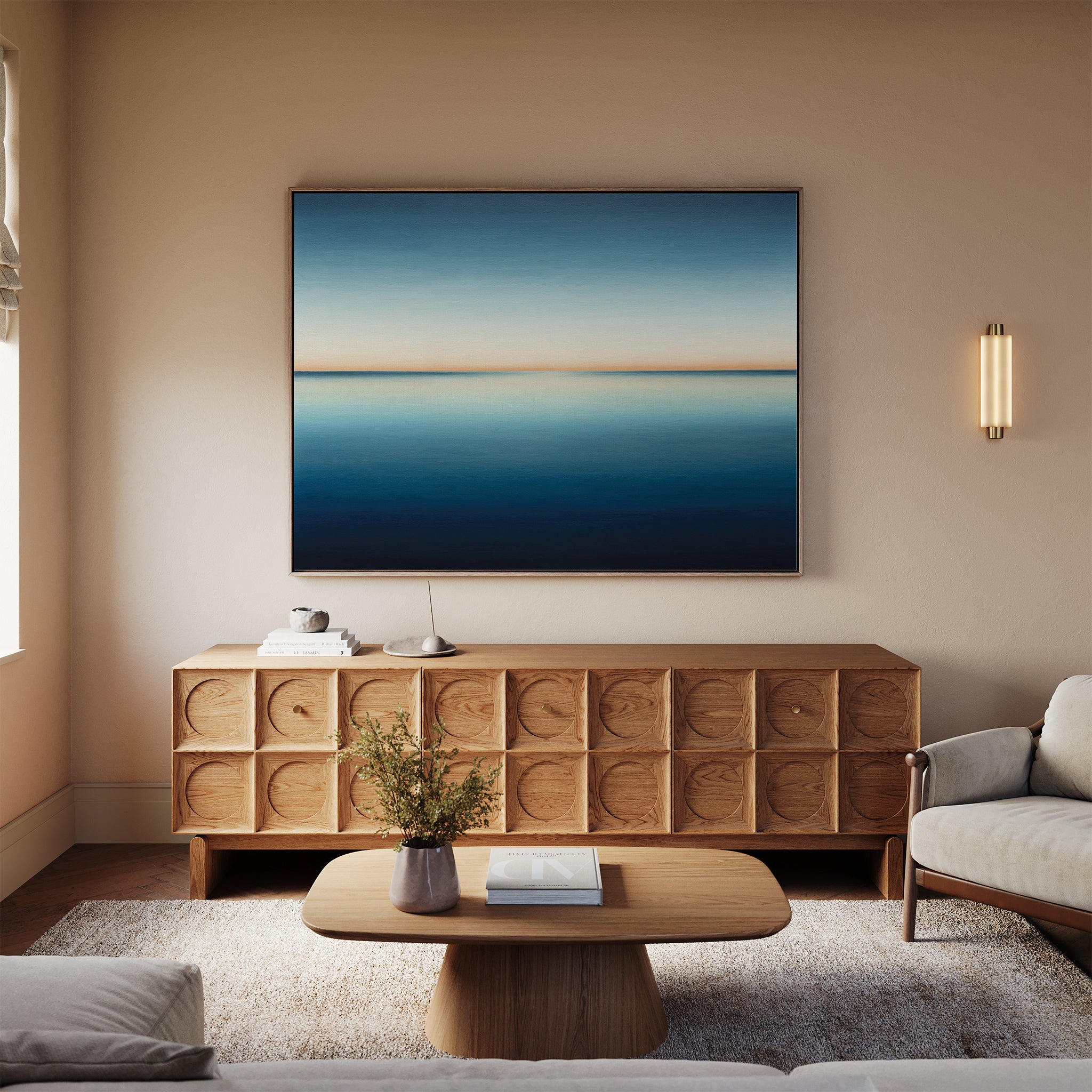 A modern living room features a wooden sideboard, the Indigo Passage Canvas Art – Horizontal in blue and beige tones, a coffee table with books, a light rug, beige sofa, wall sconce, and a small vase with flowers for stylish decor.