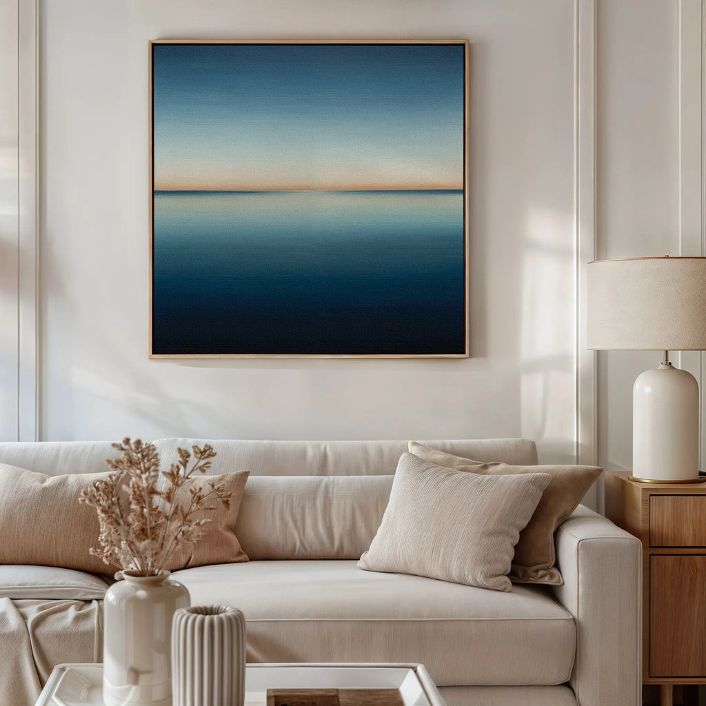 A modern living room with a beige sofa, light cushions, a side table with a ceramic lamp, dried flowers in a vase, and minimalist wall art—the Indigo Passage Canvas Art—featuring a calm blue and beige abstract horizon above the sofa.