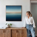 A woman in a white shirt and jeans stands by a wooden sideboard, admiring the Indigo Passage Canvas Art—a minimalist abstract seascape—framed on a white wall. Modern decor accents like a plant and books complete the serene living room scene.