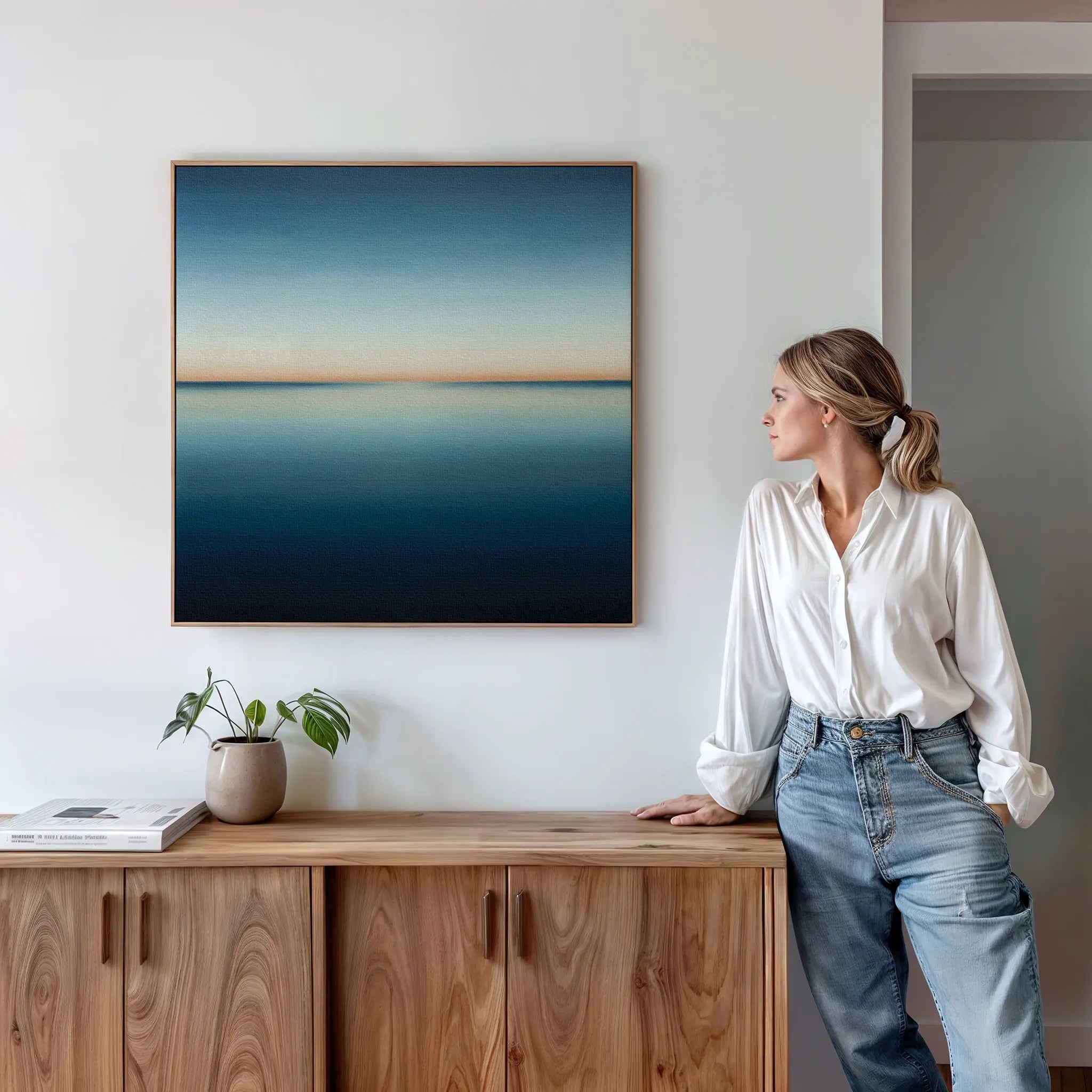 A woman in a white shirt and jeans stands by a wooden sideboard, admiring the Indigo Passage Canvas Art—a minimalist abstract seascape—framed on a white wall. Modern decor accents like a plant and books complete the serene living room scene.
