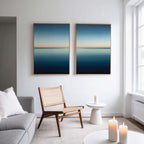 A modern living room with a grey sofa, wooden chair, small round white table, and lit candles. The Indigo Passage Set of 2 minimalist wall art in blue and gold hangs above the chair, adding a stylish touch to the decor.