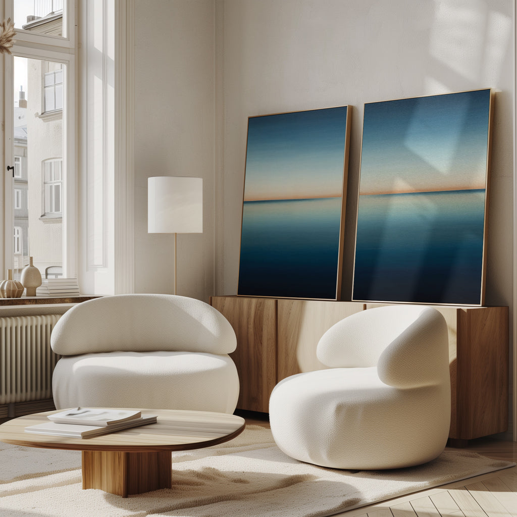 A modern, sunlit living room with two white rounded chairs, a low wooden table, and a sideboard displaying the Indigo Passage Set of 2 abstract blue seascape paintings—perfect for modern living room decor.