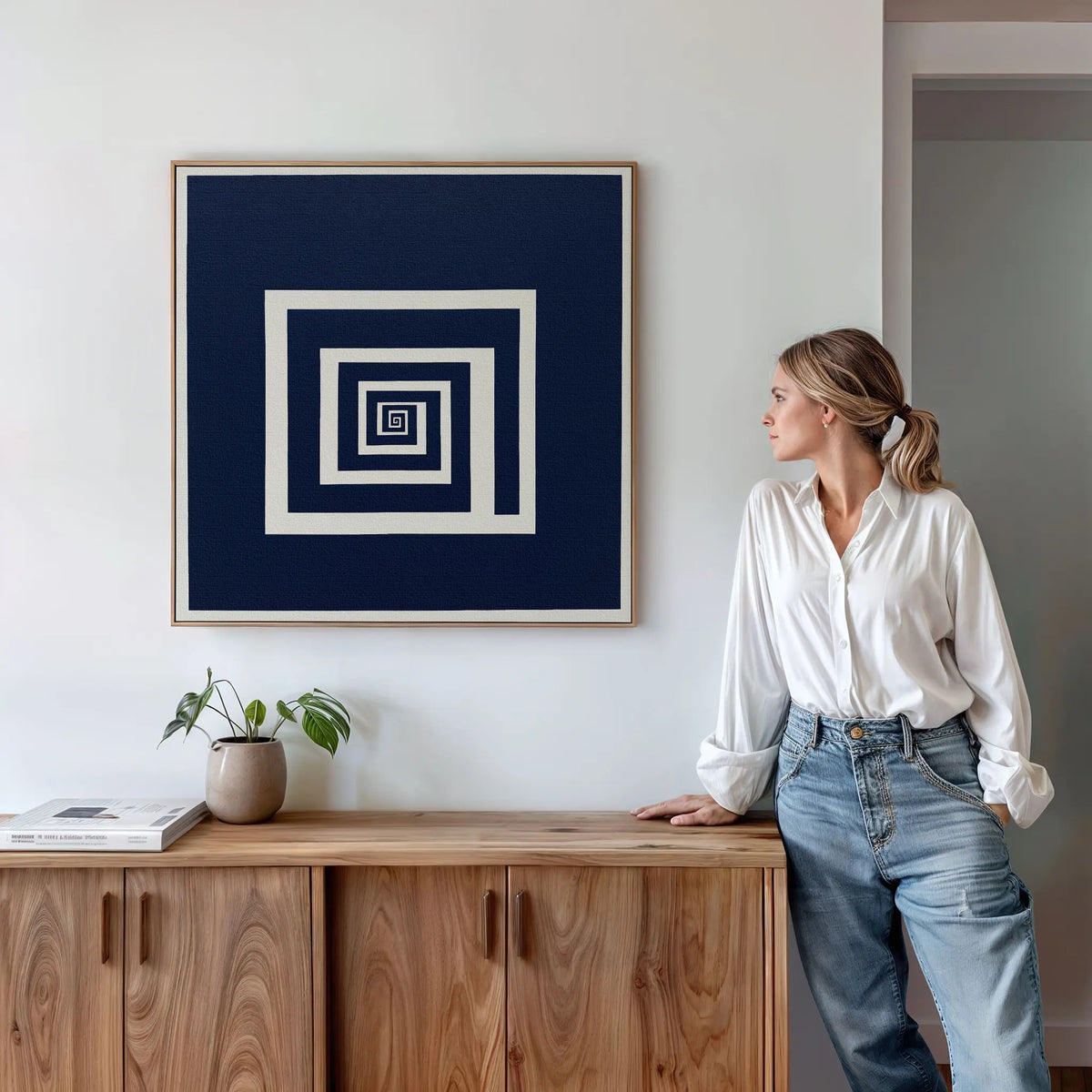 A woman in a white blouse and jeans stands by a wooden cabinet, admiring the Infinite Descent Canvas Art with navy and white geometric squares. A small potted plant and books sit on the cabinet, enhancing the modern decor.