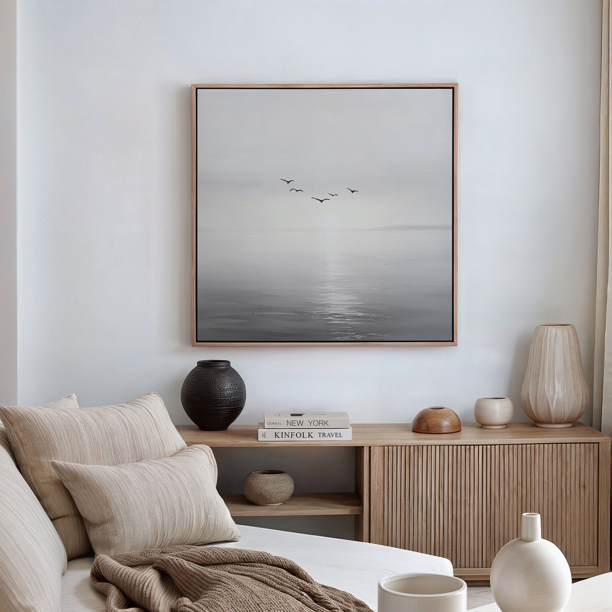 A serene, minimalist living room with a beige sofa, wooden sideboard, and neutral pillows is enhanced by the Into the Light Canvas Art depicting birds over a calm sea, adding peaceful elegance to the decor bathed in natural light.