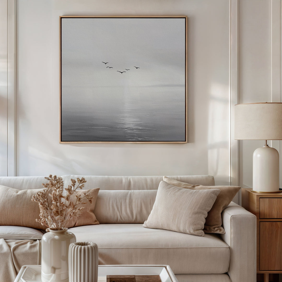 A modern living room with a white sofa, beige cushions, a side table with a cream lamp, and dried flowers. Above the sofa hangs the Into the Light Canvas Art, featuring birds over a calm gray sea—ideal for stylish decor.