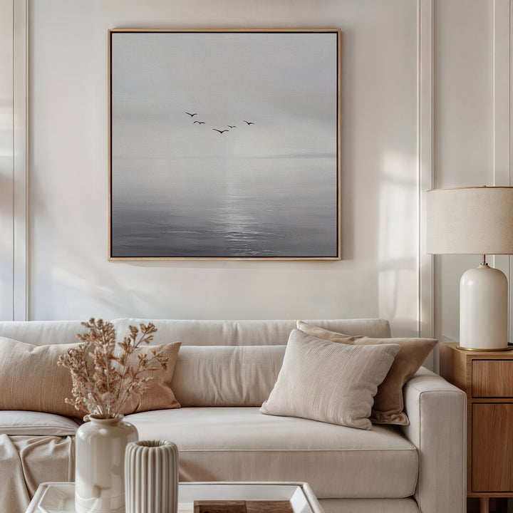 A modern living room with a white sofa, beige cushions, a side table with a cream lamp, and dried flowers. Above the sofa hangs the Into the Light Canvas Art, featuring birds over a calm gray sea—ideal for stylish decor.