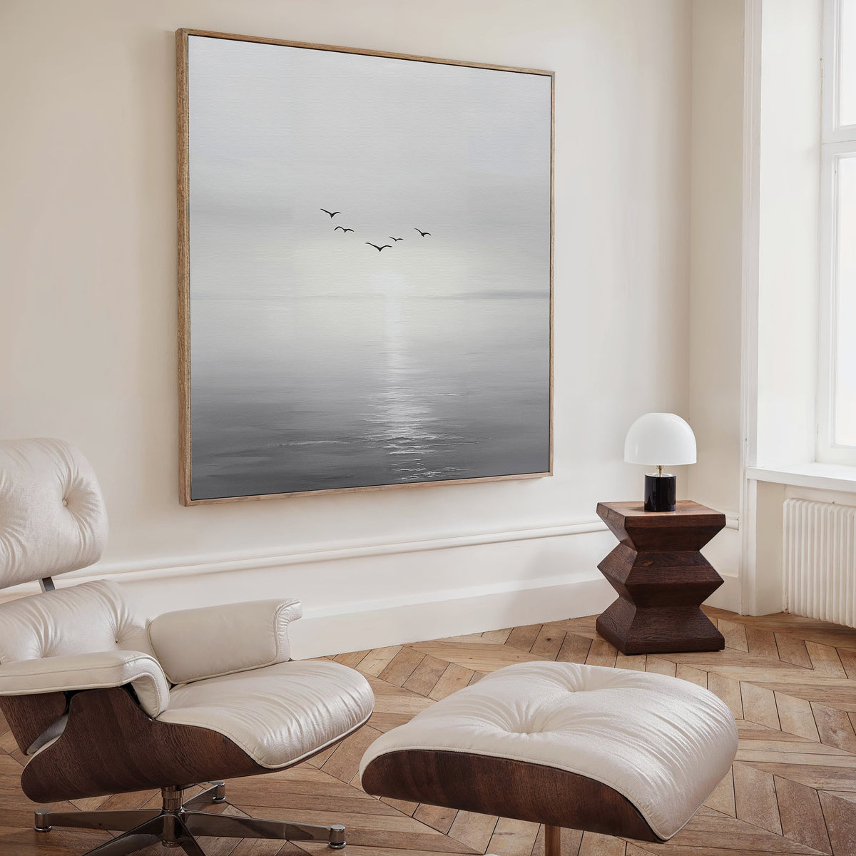 A modern, minimalist living room featuring a cream lounge chair and ottoman, wooden side table with lamp, and the striking “Into the Light Canvas Art”—birds over a misty sea at sunrise—perfect for elevated decor.