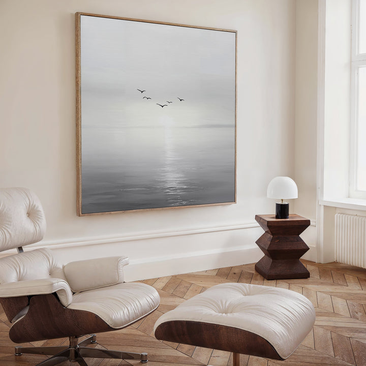 A modern, minimalist living room featuring a cream lounge chair and ottoman, wooden side table with lamp, and the striking “Into the Light Canvas Art”—birds over a misty sea at sunrise—perfect for elevated decor.
