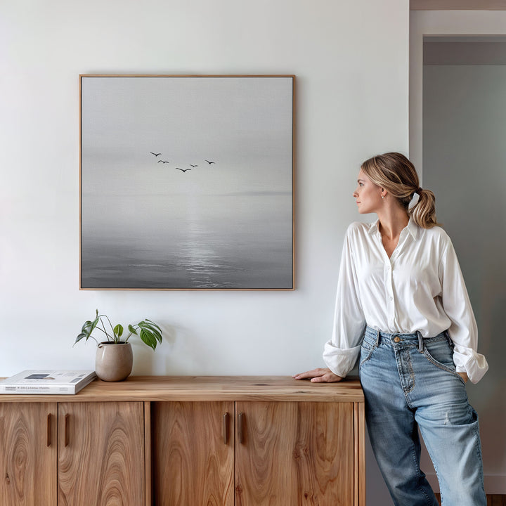 A woman in a white shirt and jeans leans against a wooden cabinet, admiring the Into the Light Canvas Art—minimalist wall art of birds flying over water at sunrise—displayed above a plant and books in her serene living room.
