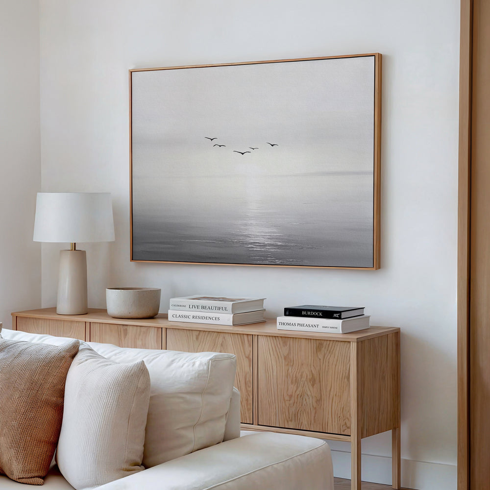 A minimalist living room features a wooden sideboard with books, a lamp, and decor accents. Above, the “Into the Light Canvas Art – Horizontal” depicts birds soaring over a calm sea in soft tones—ideal for modern decor.