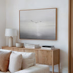 A minimalist living room features a wooden sideboard with books, a lamp, and decor accents. Above, the “Into the Light Canvas Art – Horizontal” depicts birds soaring over a calm sea in soft tones—ideal for modern decor.