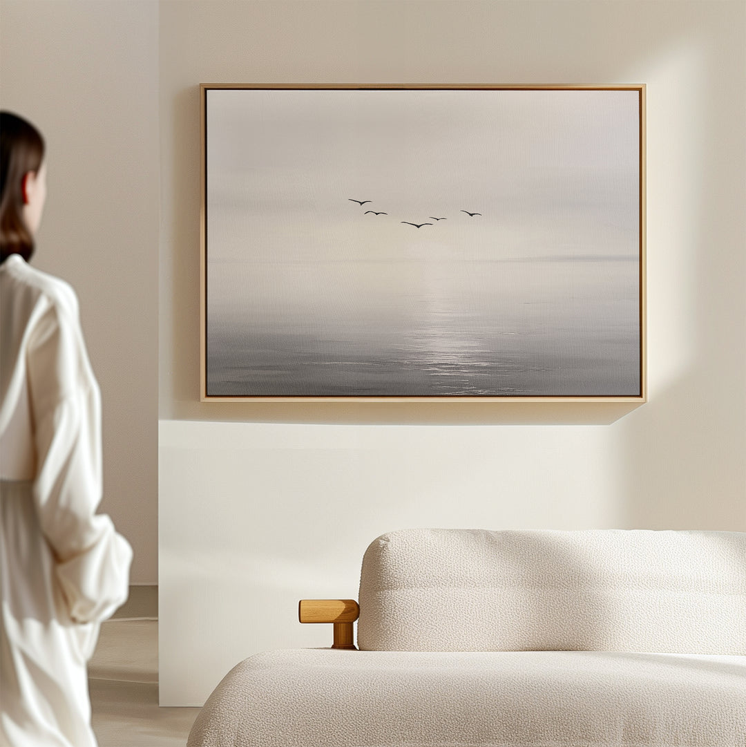 A woman in a white shirt stands in a minimal, bright room, admiring the "Into the Light Canvas Art – Horizontal" depicting birds over a misty sea, elegantly displayed above a beige upholstered sofa as stylish living room decor.