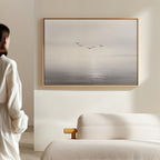 A woman in a white shirt stands in a minimal, bright room, admiring the "Into the Light Canvas Art – Horizontal" depicting birds over a misty sea, elegantly displayed above a beige upholstered sofa as stylish living room decor.