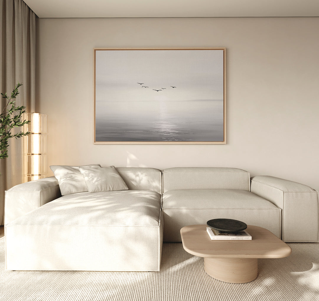 A modern living room with a beige sectional sofa, low wooden coffee table, light walls, and the "Into the Light Canvas Art – Horizontal" depicting birds over a calm sea above the sofa. A tall plant and sheer curtains are on the left.