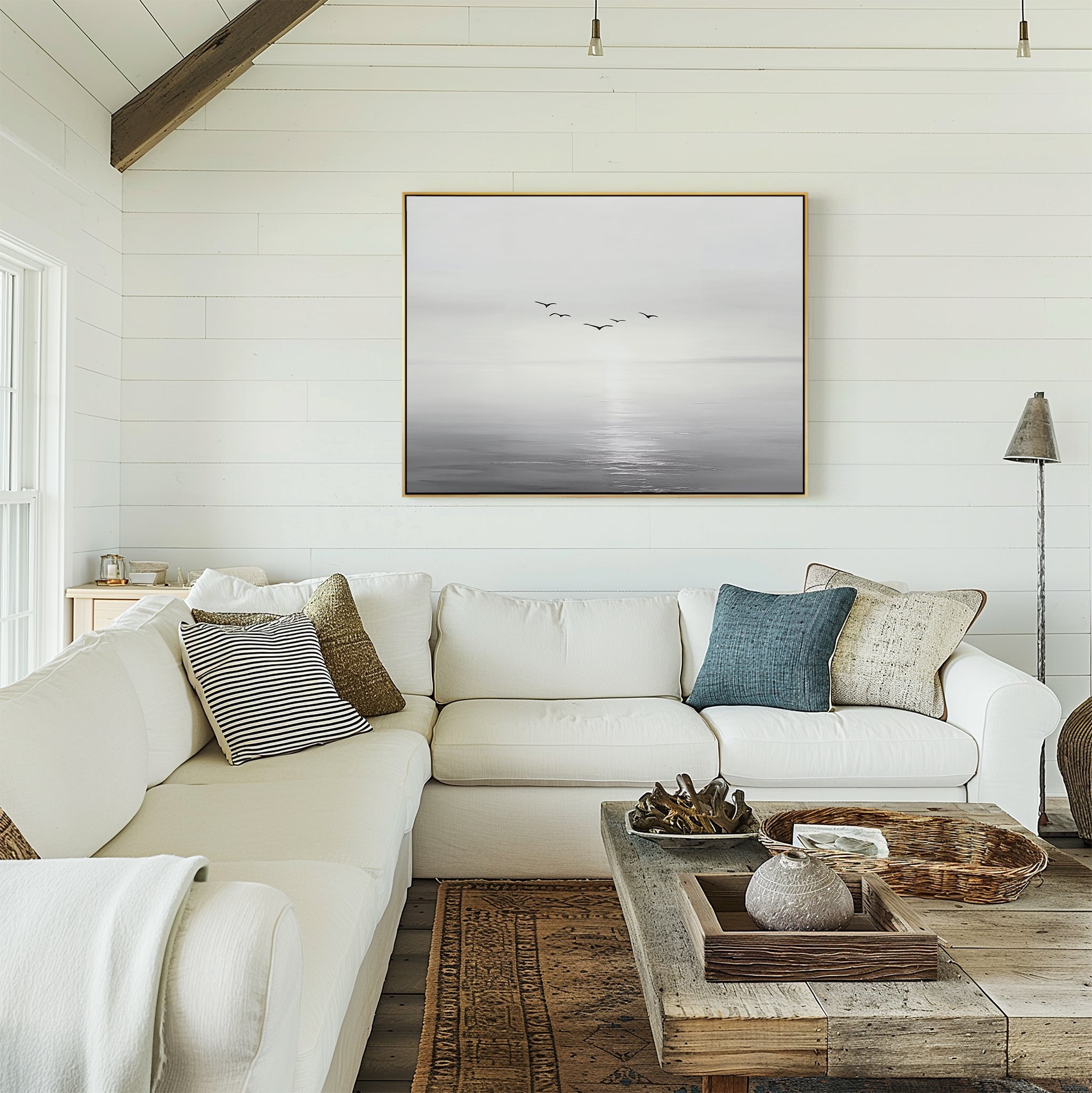 A cozy living room with white walls, a large sectional sofa, rustic wooden coffee table, assorted throw pillows, and the Into the Light Canvas Art – Horizontal—a black-and-white seascape—perfects the decor.
