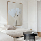 A modern living room with a cream sectional sofa, round black coffee table, and the Ivory Buds Canvas Art featuring two white flowers. Neutral tones and minimalist decor create a calm, elegant atmosphere.