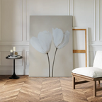 A minimalist room features the Ivory Buds Canvas Art—two white flowers on canvas—leaning against the wall, with an empty frame behind, a round black table with candles and books, and a white cushioned chair on herringbone floors.