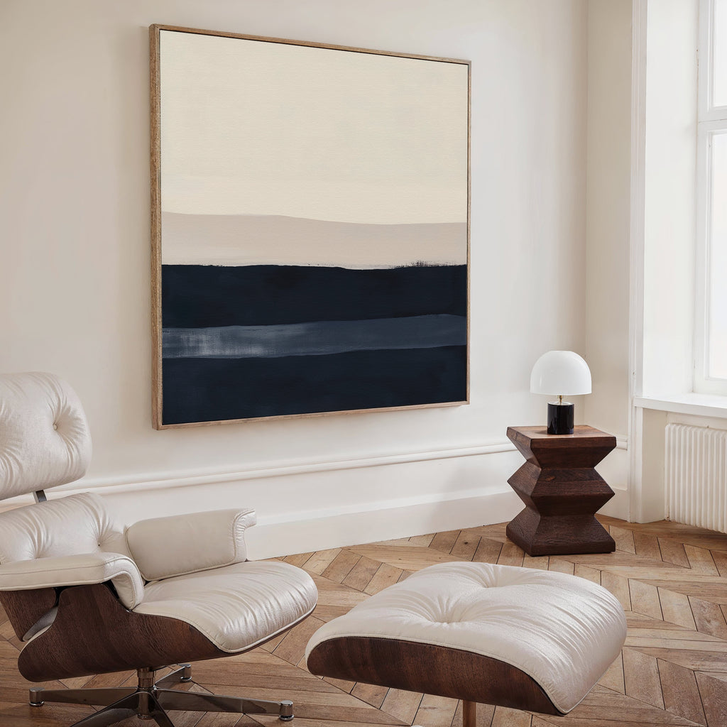 A modern, minimalist living space with a white Eames lounge chair and ottoman, a geometric wooden side table topped with a white lamp, and the Ivory Dusk Canvas Art featuring beige and navy bands—ideal for fans of modern interiors.