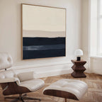 A modern, minimalist living space with a white Eames lounge chair and ottoman, a geometric wooden side table topped with a white lamp, and the Ivory Dusk Canvas Art featuring beige and navy bands—ideal for fans of modern interiors.