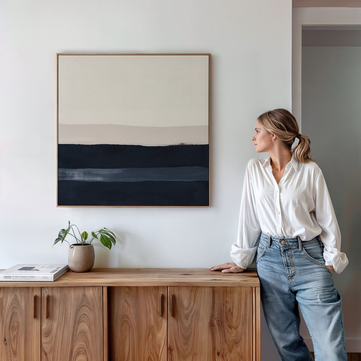 A woman in a white shirt and jeans stands by a wooden cabinet, admiring the Ivory Dusk Canvas Art featuring horizontal bands of beige, tan, and dark blue. Perfect for modern interiors, its styled with a small plant and books on the cabinet.