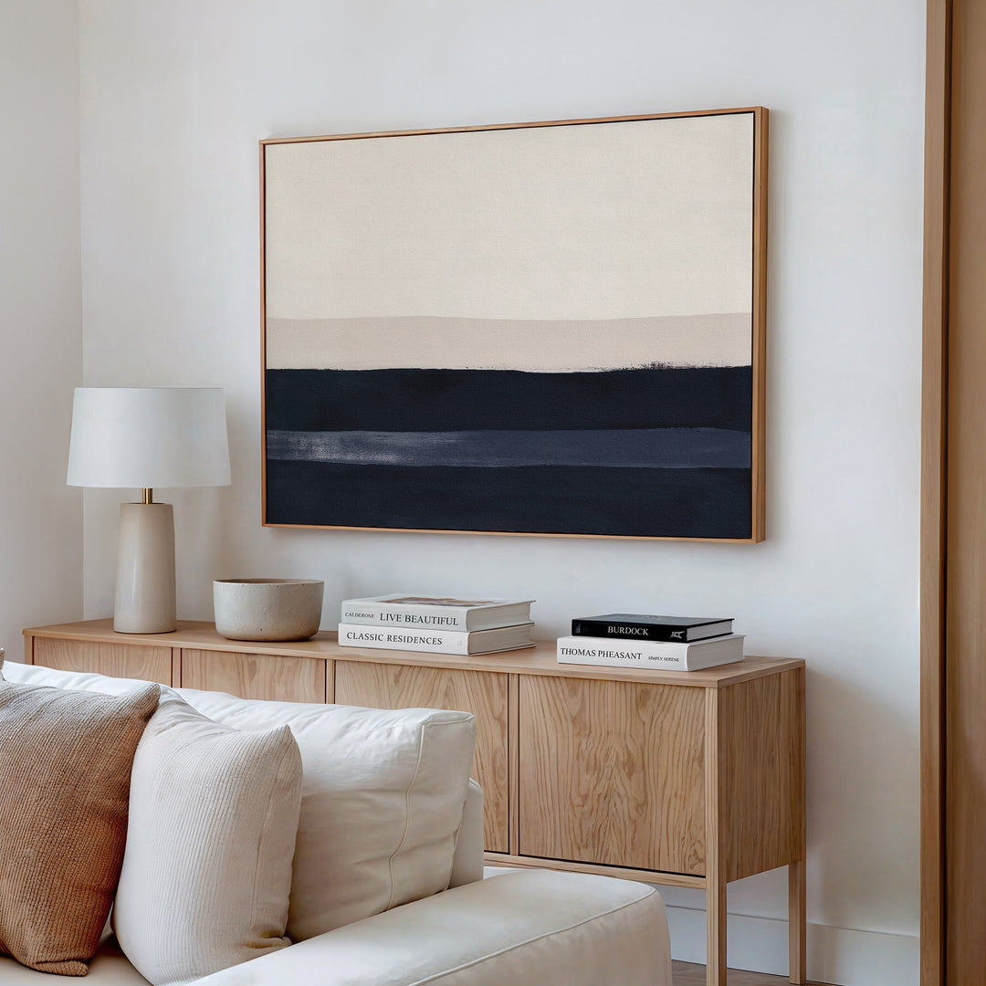A modern, minimalist living room features a wooden sideboard with books, a bowl, and a lamp, a beige sofa, and the Ivory Dusk Canvas Art – Horizontal in neutral tones hanging on the white wall above the sideboard.