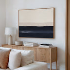 A modern, minimalist living room features a wooden sideboard with books, a bowl, and a lamp, a beige sofa, and the Ivory Dusk Canvas Art – Horizontal in neutral tones hanging on the white wall above the sideboard.