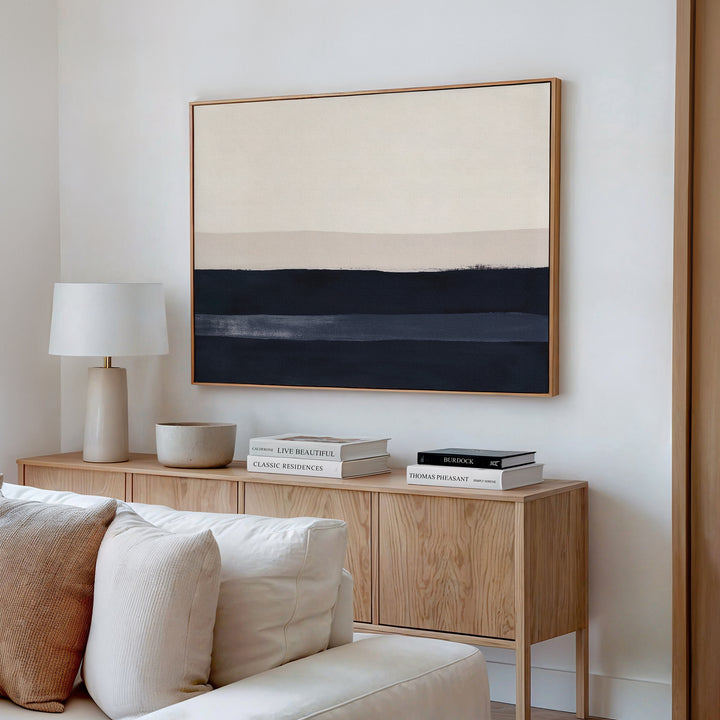 A modern, minimalist living room features a wooden sideboard with books, a bowl, and a lamp, a beige sofa, and the Ivory Dusk Canvas Art – Horizontal in neutral tones hanging on the white wall above the sideboard.
