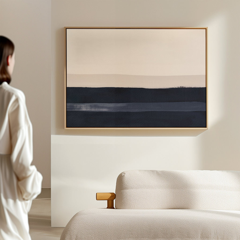 A person in a white outfit stands by a minimalist living room with a light sofa and the Ivory Dusk Canvas Art – Horizontal, featuring bold beige and dark blue bands as striking modern wall decor.