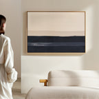 A person in a white outfit stands by a minimalist living room with a light sofa and the Ivory Dusk Canvas Art – Horizontal, featuring bold beige and dark blue bands as striking modern wall decor.
