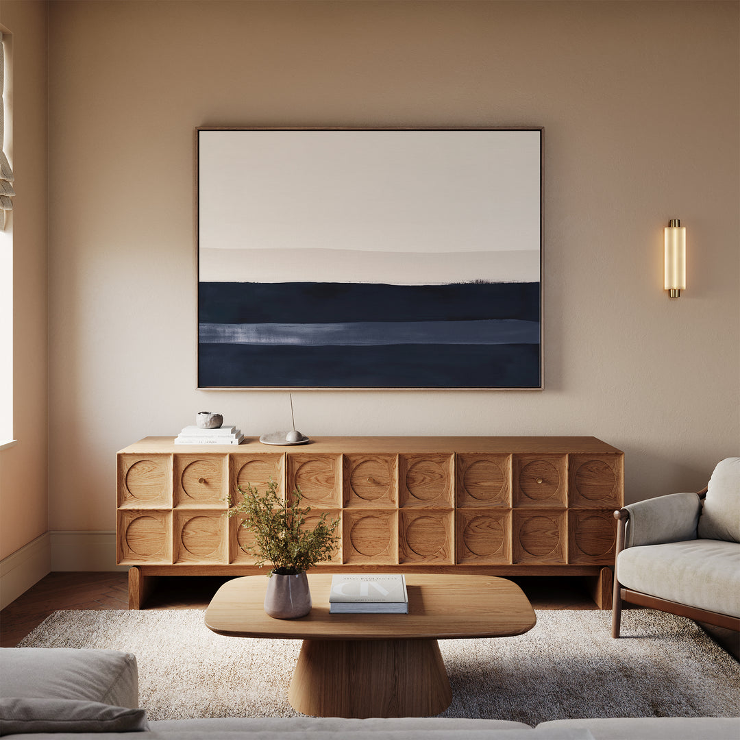 A modern living room features a geometric wooden sideboard, a low wooden coffee table, a light gray sofa, a textured rug, and the Ivory Dusk Canvas Art – Horizontal in neutral and dark blue tones for a sophisticated touch.